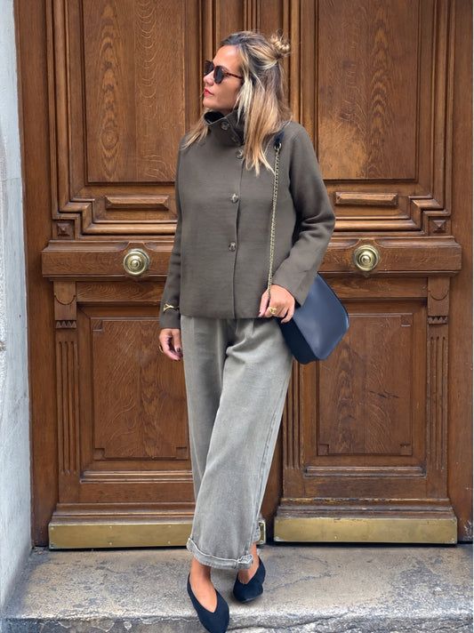 Gilet Pietra MYLO kaki à col montant porté en pied avec un pantalon clair devant une porte en bois